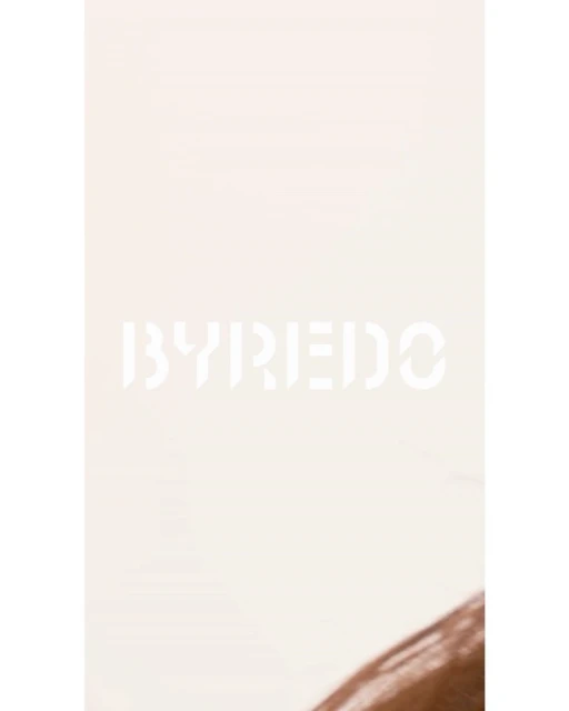 Close-up studio shot of light brown hair with dark roots, styled with a slight wave, against a beige background with the 'BYREDO' logo.