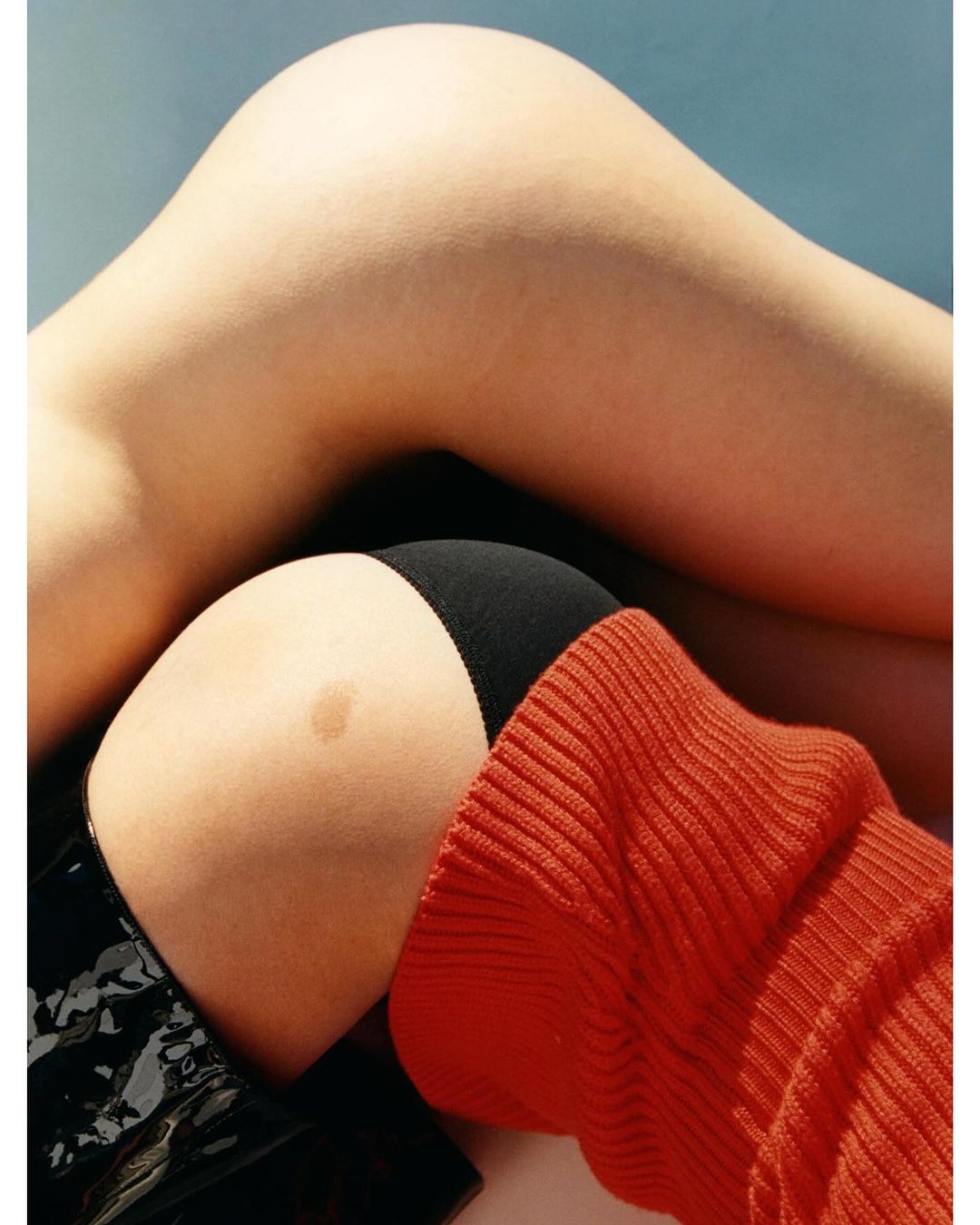 Close-up studio shot of a person's legs and torso with black underwear, a bright orange sweater, and a shiny black coat.