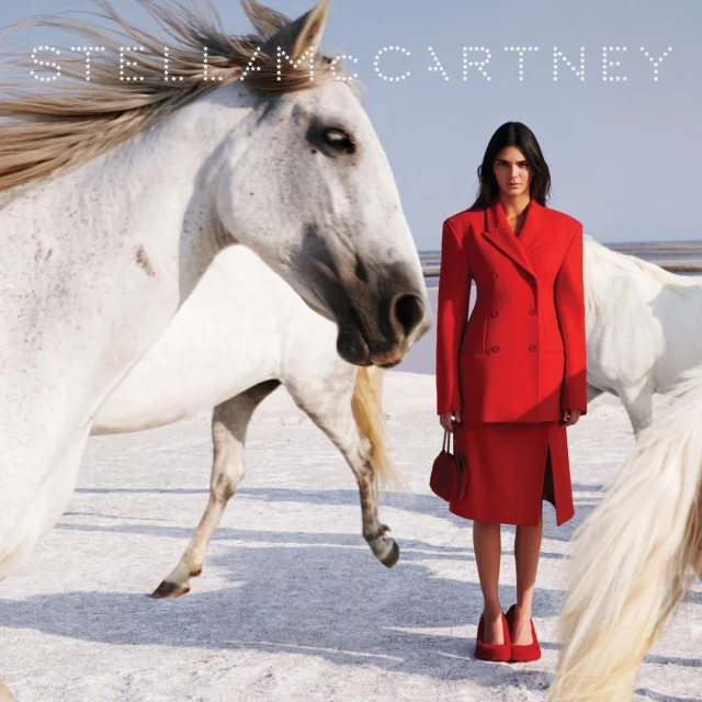 Fashion editorial featuring a model in a red suit standing among white horses on a salt flat under a bright sky for Stella McCartney.