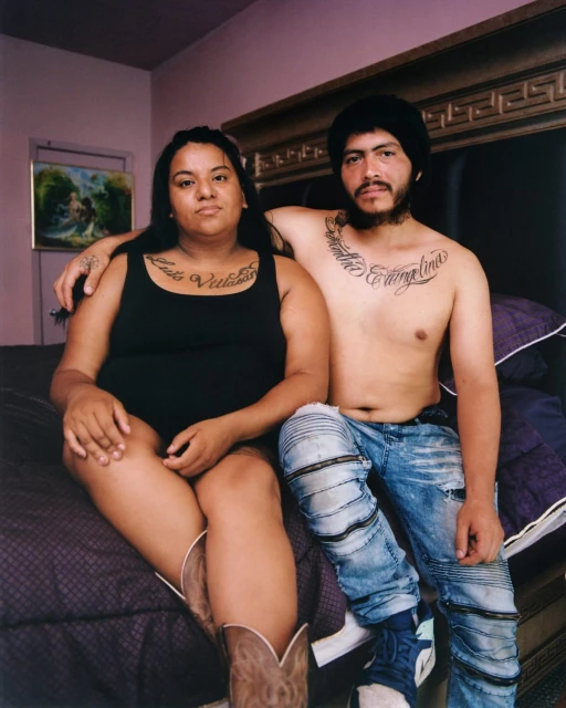 An intimate portrait of a Hispanic couple sitting on a bed in a room, featuring tattoos, cowboy boots, and casual attire in a candid, lifestyle shot.