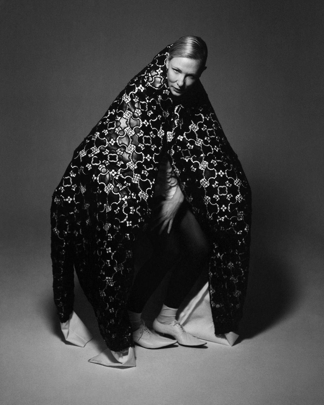 Monochrome portrait of a woman enveloped in a patterned lace covering against a grey backdrop, showcasing fashion and studio photography.
