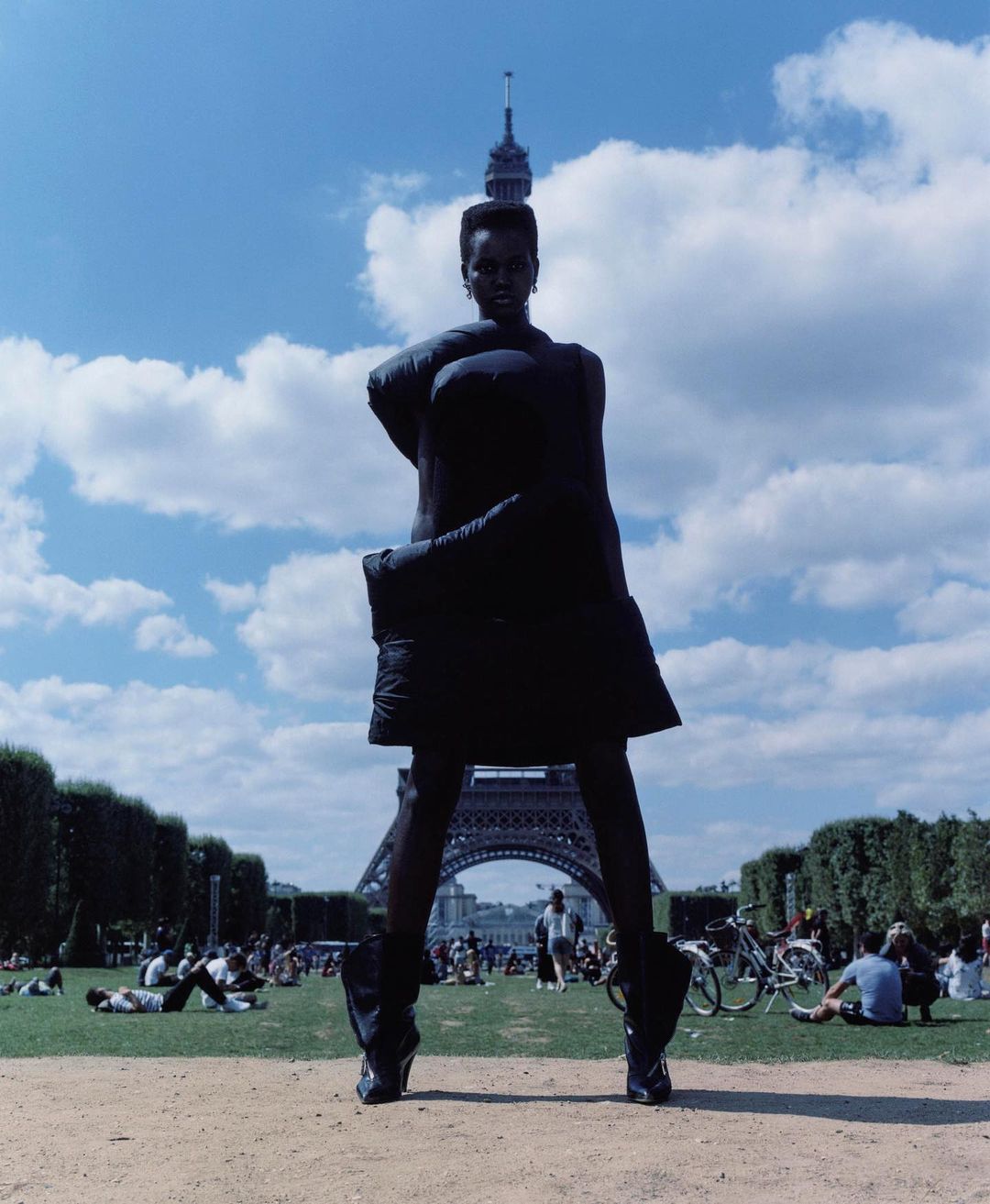 Model standing outdoors with arms wrapped around torso in front of the Eiffel Tower under a blue sky with scattered clouds.