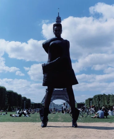Model standing outdoors with arms wrapped around torso in front of the Eiffel Tower under a blue sky with scattered clouds.