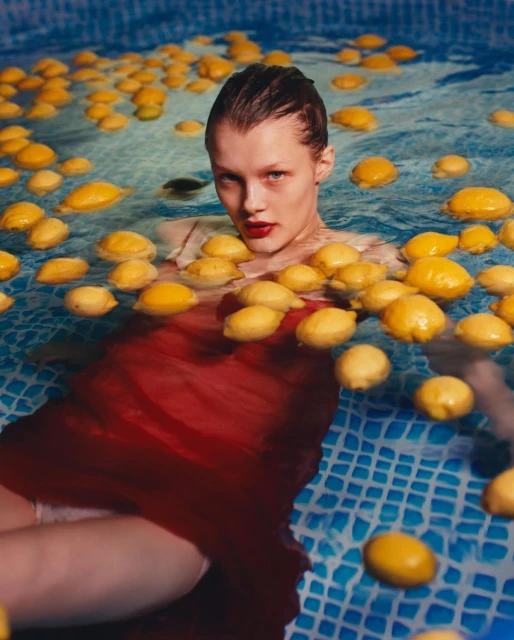 Woman in red dress partially submerged in a blue-tiled pool filled with floating lemons, gazing directly at the camera.