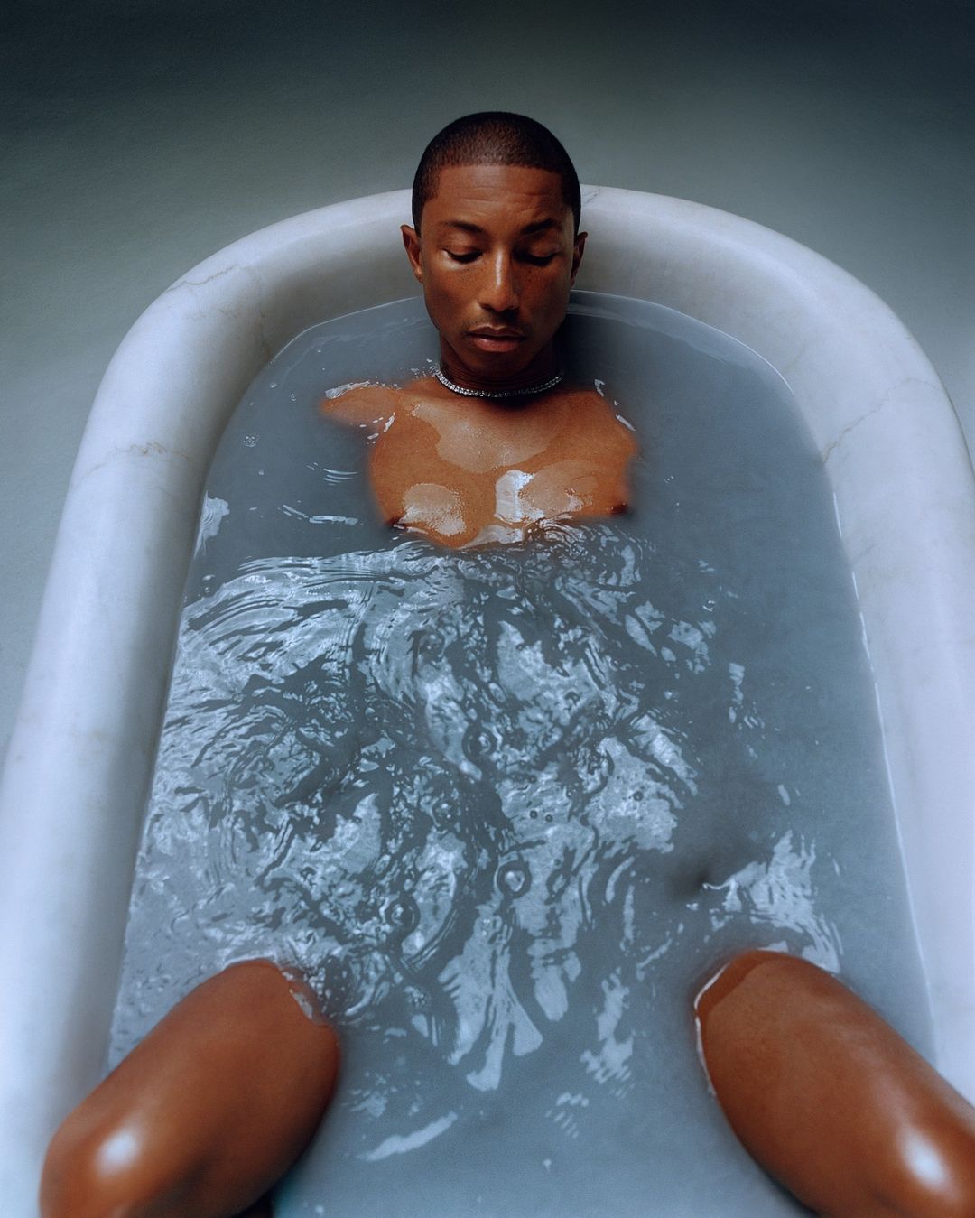 Adult male musician, Pharrell Williams, poses in a marble bathtub filled with water, wearing a diamond necklace, for a portrait.