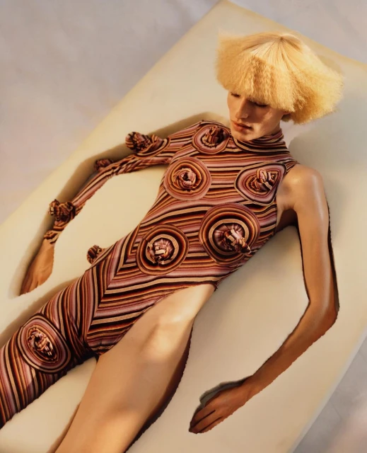 Woman with blond crimped hair lies on a padded surface wearing a patterned bodysuit with hand-shaped embellishments in a studio setting.