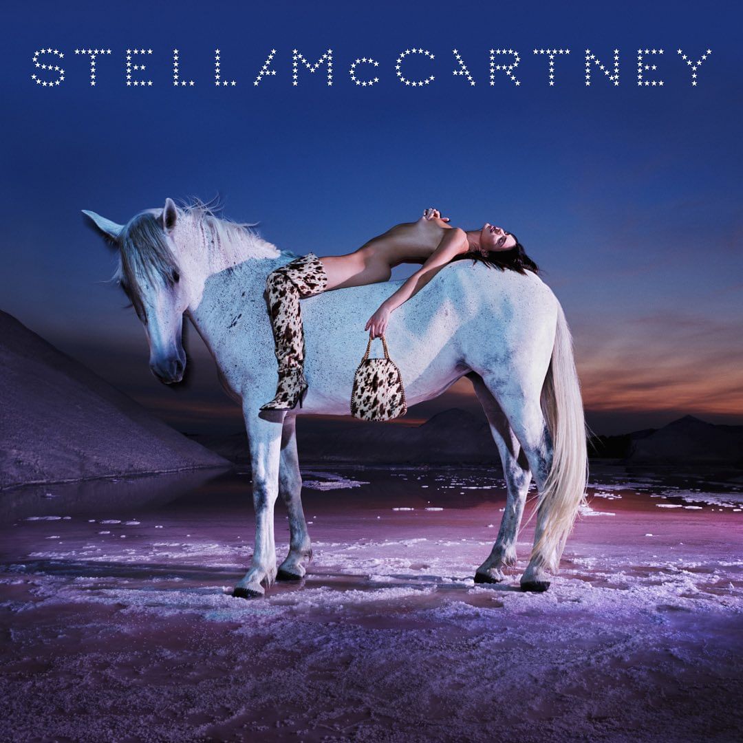 Fashion image of a topless female model with boots and a handbag lying on a white horse near a body of water in an outdoor setting.