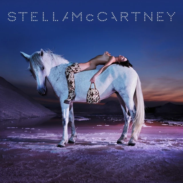 Fashion image of a topless female model with boots and a handbag lying on a white horse near a body of water in an outdoor setting.