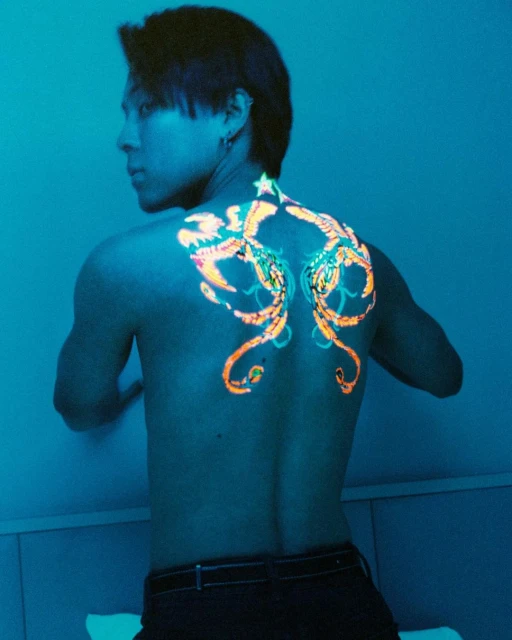 Shirtless man with back tattoos in blue light. Back turned toward viewer. The tattoos are orange and turquoise.