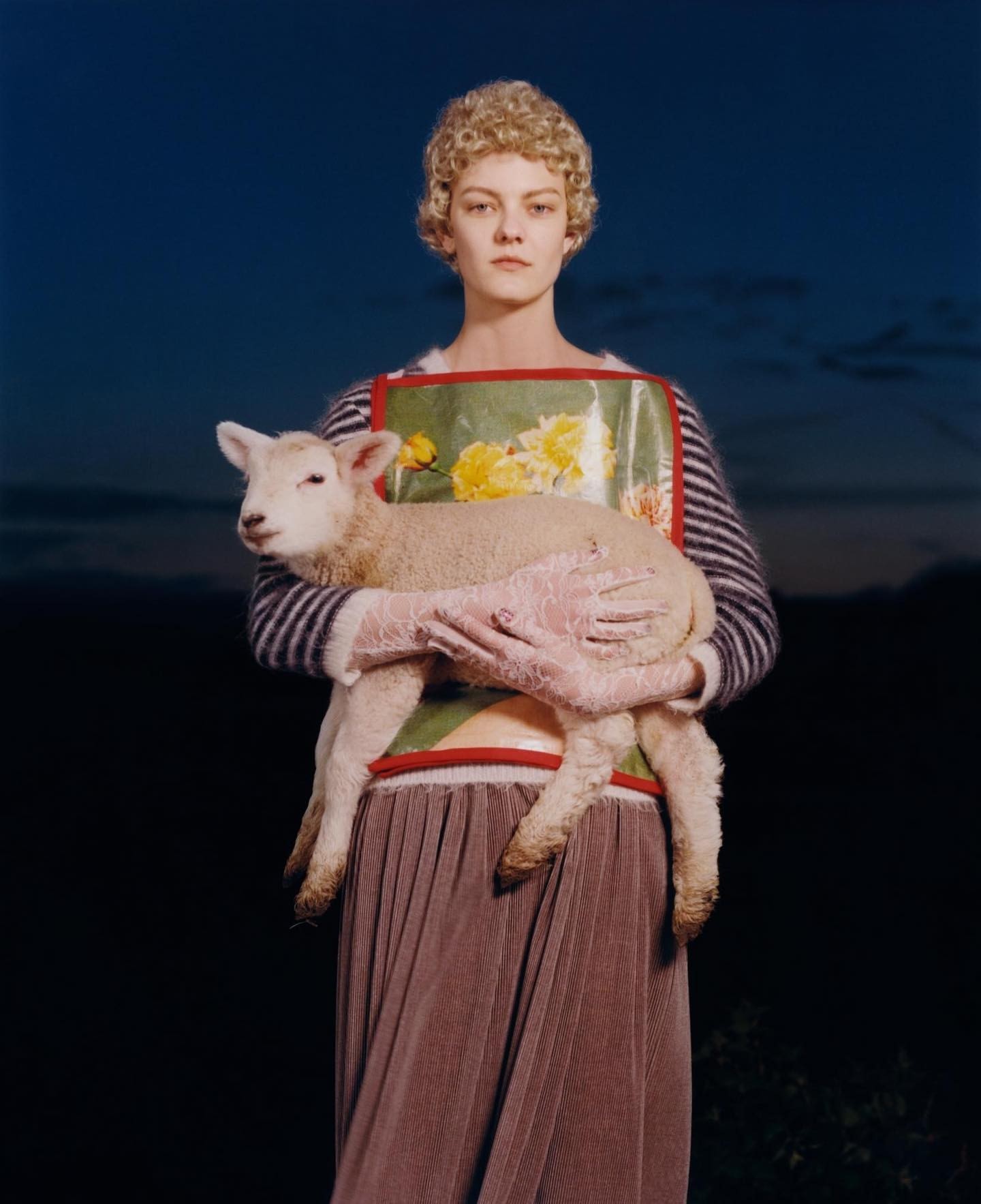 A young adult female holds a lamb against a dusky sky backdrop. She is wearing a floral apron, striped top, and pleated skirt.