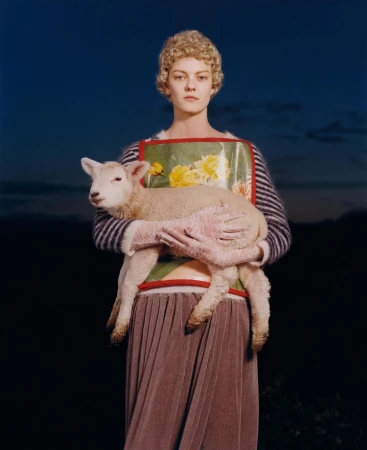 A young adult female holds a lamb against a dusky sky backdrop. She is wearing a floral apron, striped top, and pleated skirt.