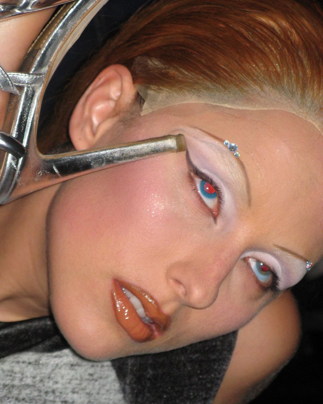 Close-up of a woman with light skin, red hair, dramatic makeup, and silver strappy heel next to her face in a portrait shot.