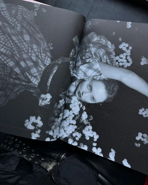 Monochrome image of a young fair-skinned woman in a patterned dress lying on a surface covered in flowers in an open book spread.