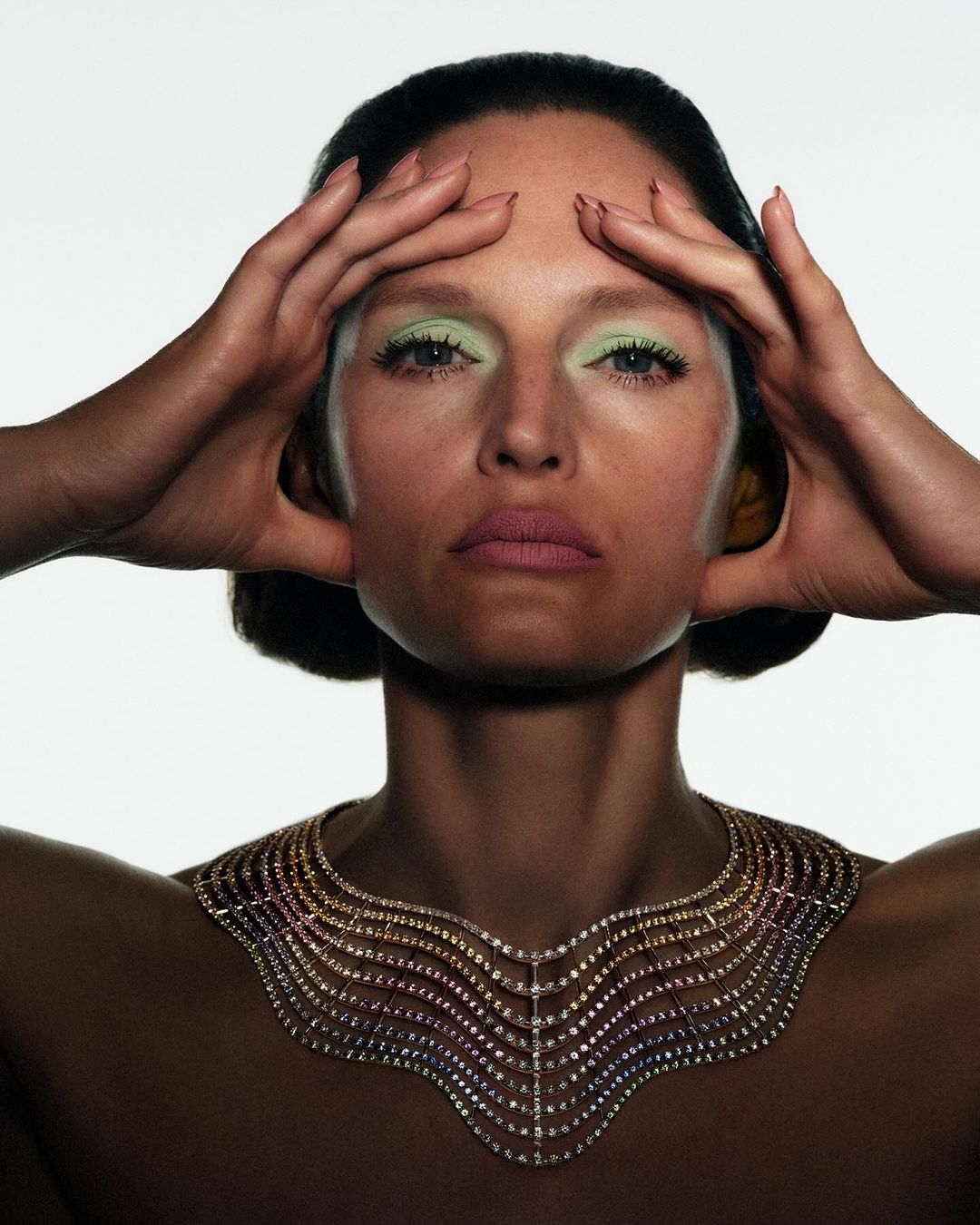 Close-up shot of a woman with light green eyeshadow and a diamond necklace, holding her hands on her head, set against a white background.