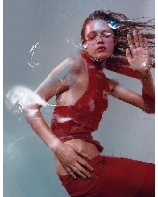Fashion shot of a female model in a red halter top and pants posing in front of a translucent surface with water droplets.