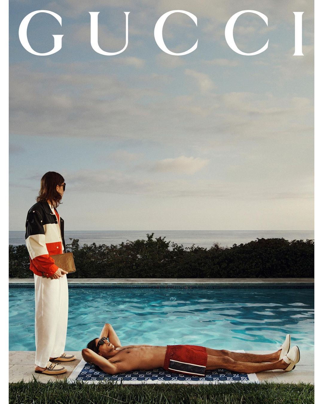 Two adults in swimwear relax poolside on a sunny day; one lies on a patterned towel, the other is stands nearby wearing a jacket.
