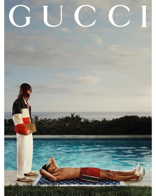 Two adults in swimwear relax poolside on a sunny day; one lies on a patterned towel, the other is stands nearby wearing a jacket.