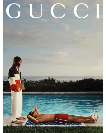 Two adults in swimwear relax poolside on a sunny day; one lies on a patterned towel, the other is stands nearby wearing a jacket.