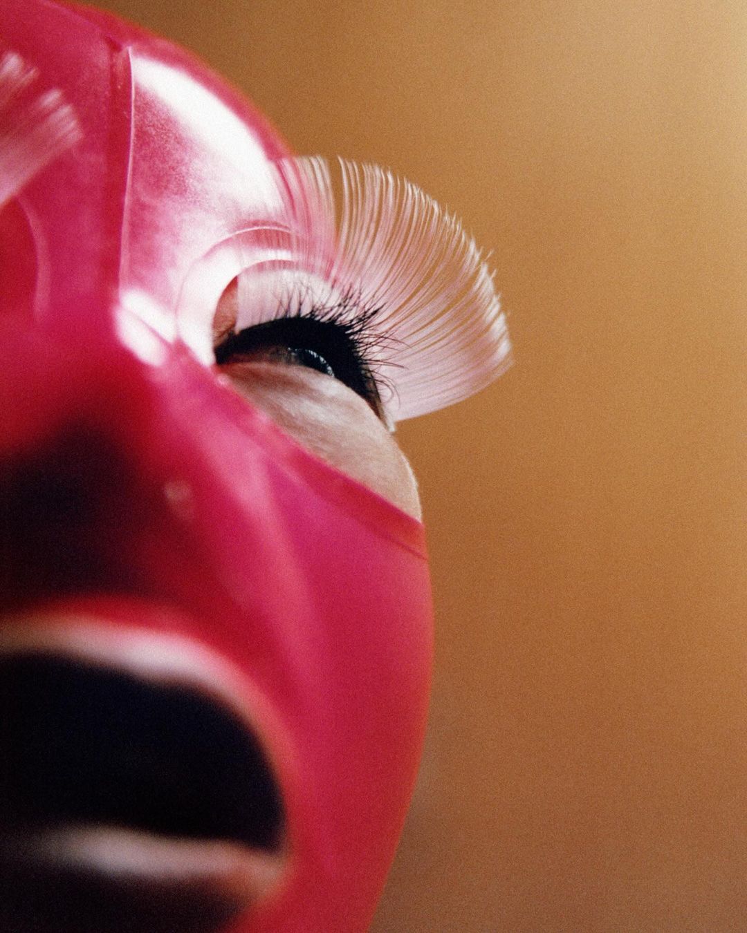 Close-up studio shot of a person wearing a bright pink latex hood, featuring dramatic white eyelashes on their right eye, against an amber background.