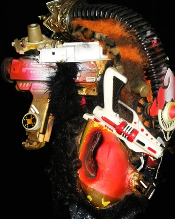 A bizarre mask-like sculpture or costume headpiece with layers of textures, materials, and toy gun attachments on a dark background.