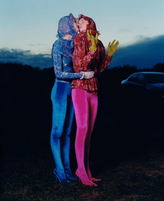 Two women embrace outdoors at dusk, wearing matching floral tops, gloves and head coverings, with one in pink leggings and the other in blue.