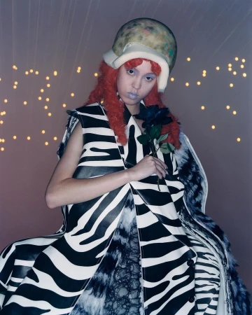 A woman with red hair and a unique helmet poses with a black rose, wearing a zebra-striped coat in a softly lit studio environment.