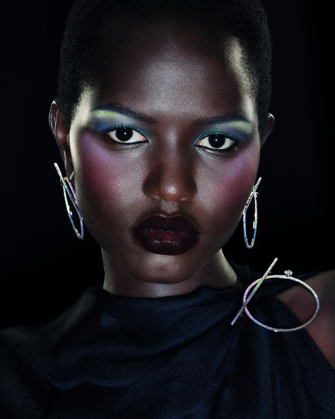 Close-up beauty shot of a striking dark-skinned woman with vibrant makeup and jewelry against a black background. Fashion and glamour.