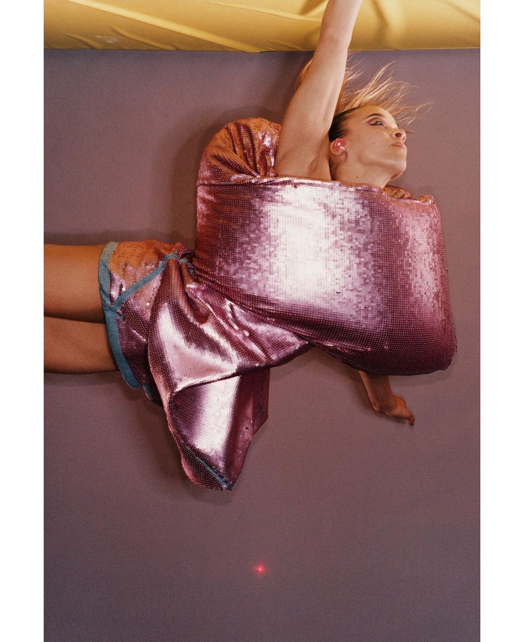 Stylish adult female fashion model in a pink sequined dress poses dynamically against a mauve backdrop with yellow lighting.