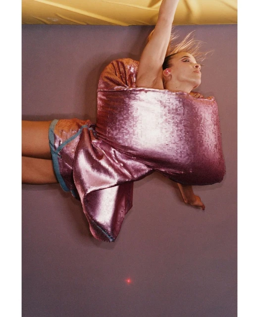 Stylish adult female fashion model in a pink sequined dress poses dynamically against a mauve backdrop with yellow lighting.