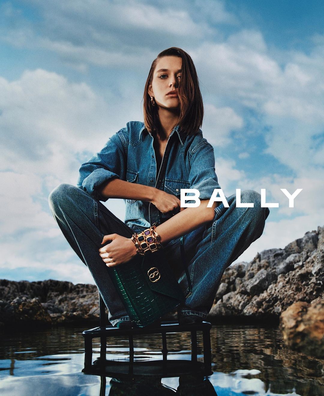 A woman in denim sits on a stool by the water, modeling a green handbag and jeweled bracelet, against a backdrop of blue sky and rocks.