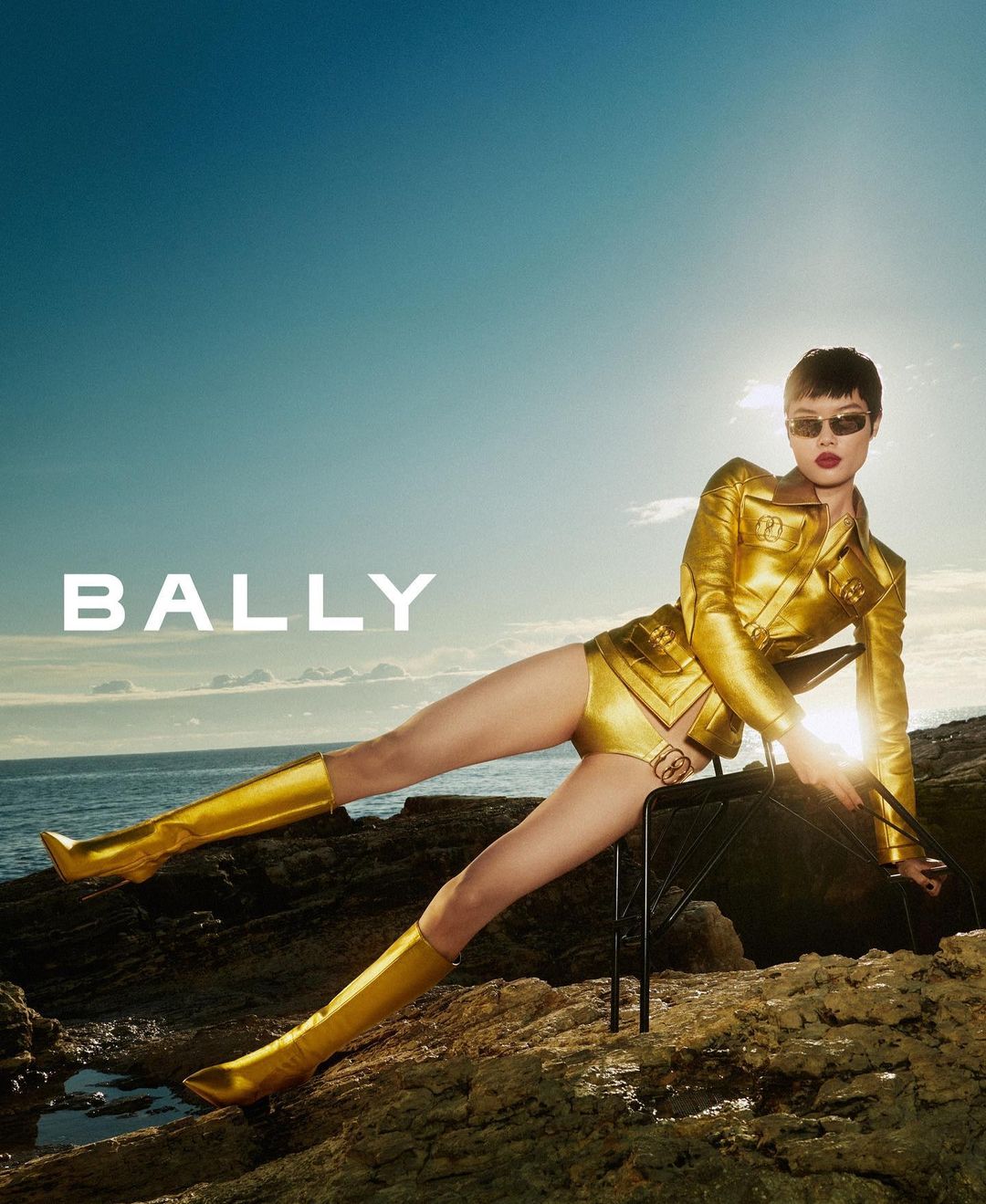 Fashion shot of a woman in a gold jacket, shorts, and boots posing on a rocky shoreline against a blue sky. Bally logo visible.