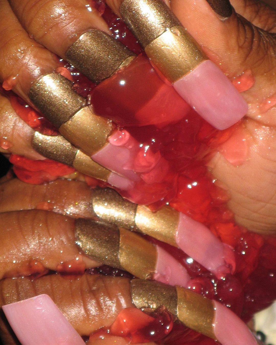 Close-up of hands with pink and gold nails immersed in red jelly, showcasing beauty and nail art in a studio setting.