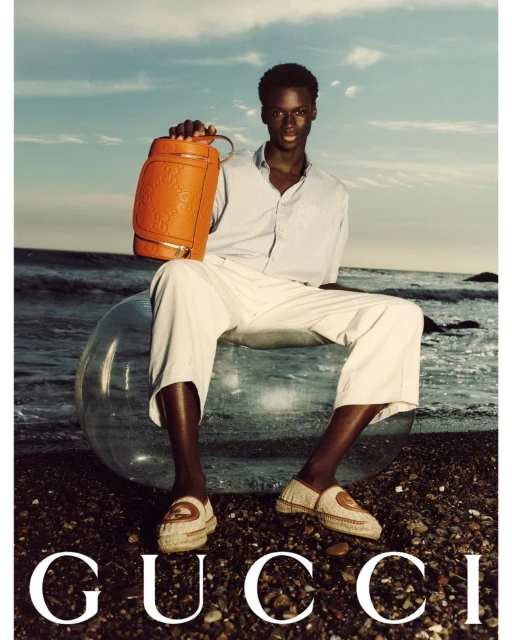 A dark-skinned male model holds a brown leather duffel bag while sitting on a translucent inflatable ball at the beach with 'GUCCI' text overlay.