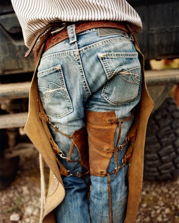 Rear view of a person wearing jeans and leather chaps, standing in front of a vehicle. Clothing details and outdoor setting visible.