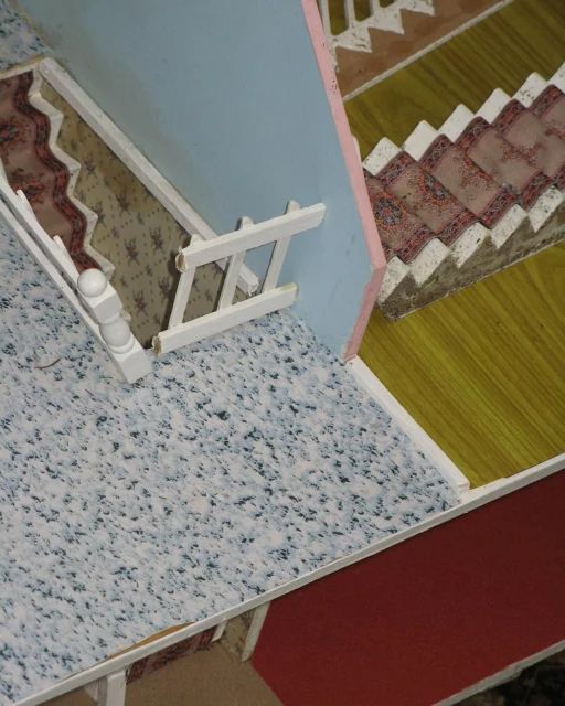 Interior view of a dollhouse featuring a staircase with patterned carpet, blue walls, and white trim, evoking a miniature, whimsical scene.