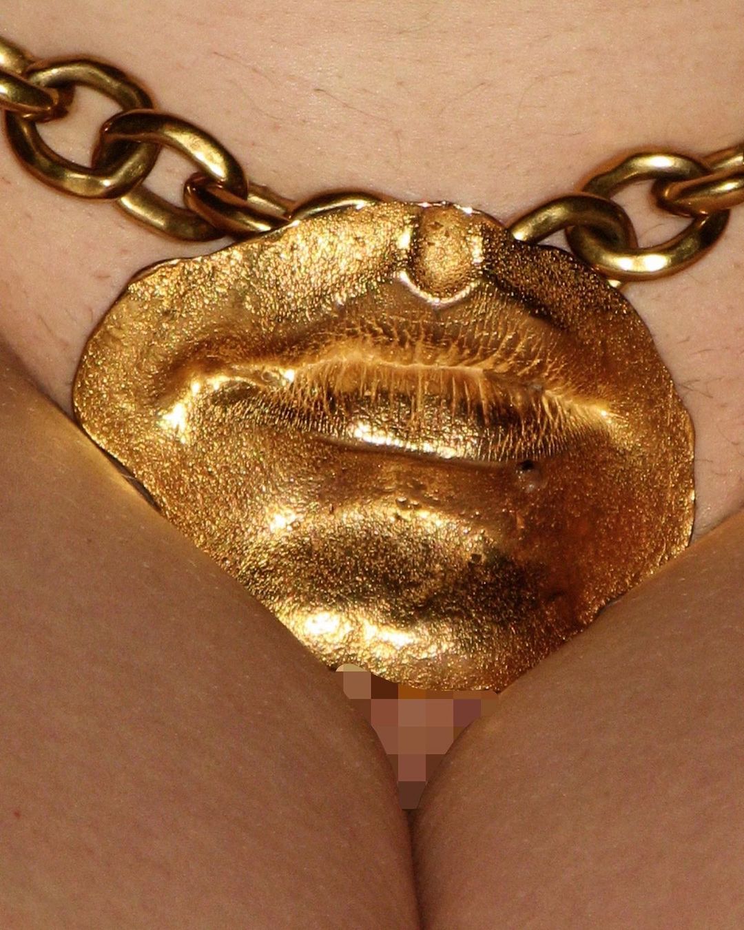 Close-up of a person wearing a gold chain necklace with a large lips-shaped pendant, partially obscuring a nude body.