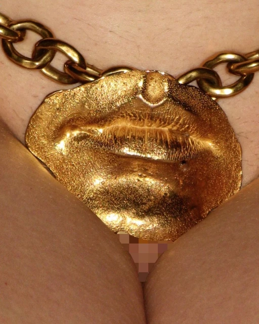 Close-up of a person wearing a gold chain necklace with a large lips-shaped pendant, partially obscuring a nude body.