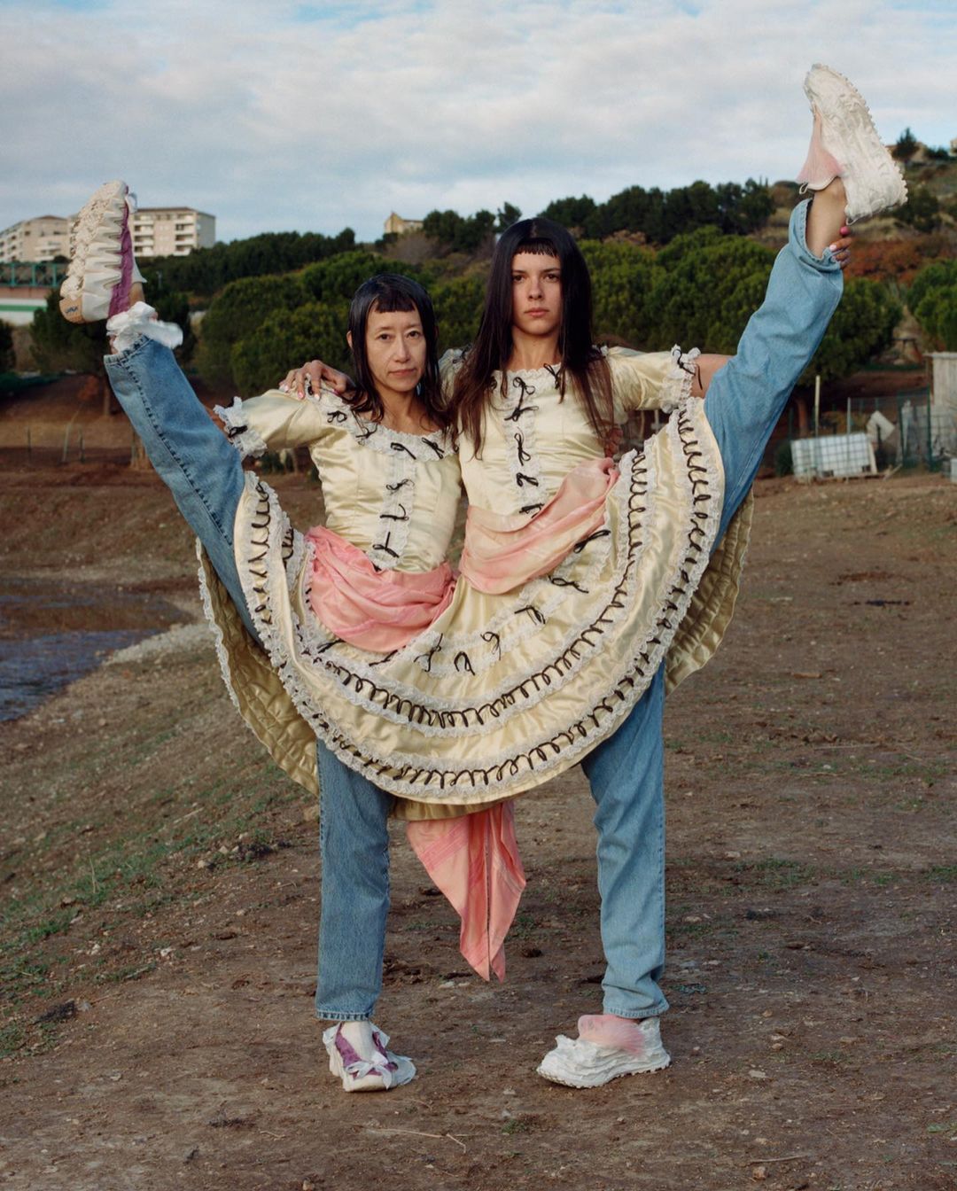 Two women in dresses and jeans pose with legs raised outdoors against a backdrop of sparse trees and a light sky, creating a quirky fashion tableau.