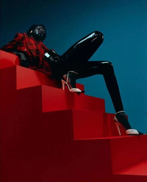 Fashion editorial shot of a Black woman posing in glossy leggings, heels, and a plaid blazer on red steps against a blue backdrop.