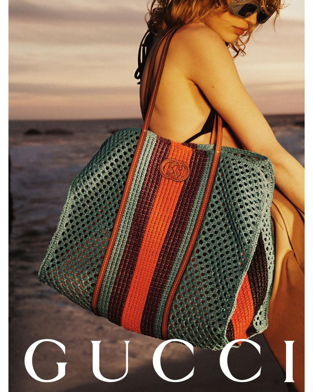 Woman wearing sunglasses and a bikini top holding a large tote bag on the beach at sunset, fashion advertisement for Gucci.