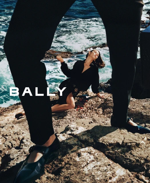 Fashion shot on rocky shore with a woman in a dark dress, foregrounded by a man's legs in dark pants and shoes and the brand 'Bally'.