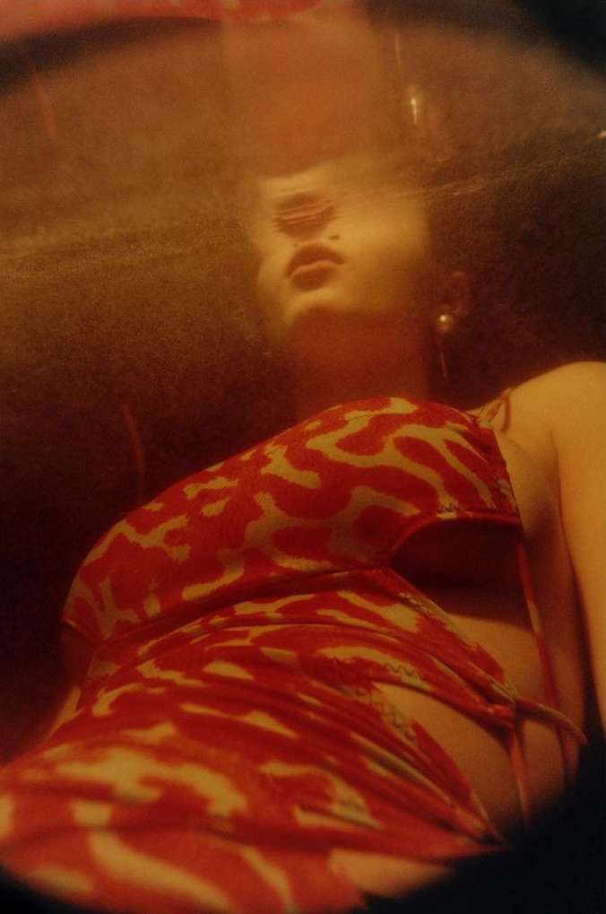 Woman with dark curly hair, looking up, wearing a red-and-white animal-print dress, in a dimly lit indoor environment with warm tones.