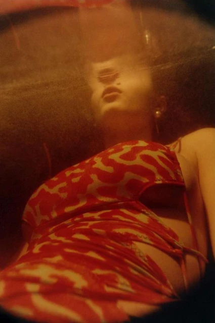 Woman with dark curly hair, looking up, wearing a red-and-white animal-print dress, in a dimly lit indoor environment with warm tones.