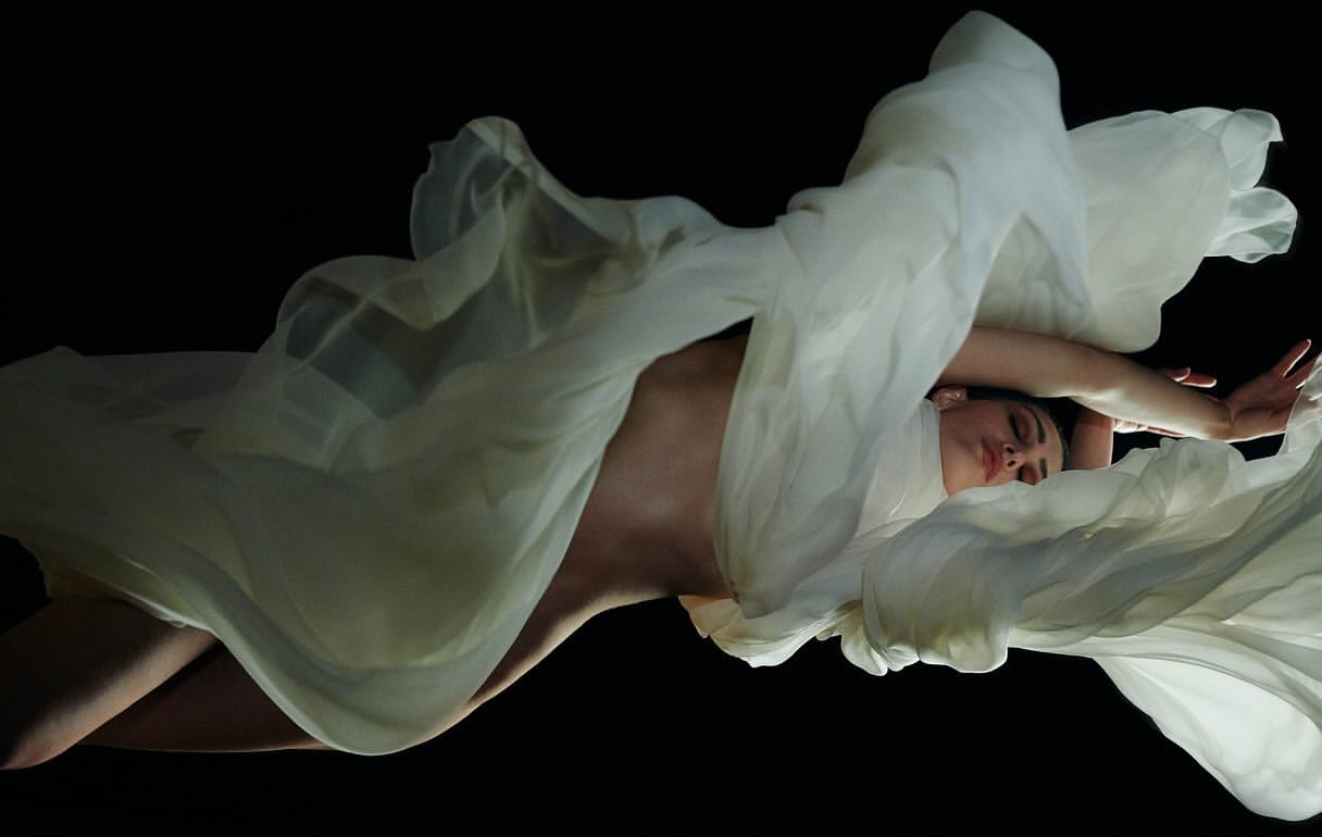 Woman draped in flowing white fabric against a black background, creating an ethereal and artistic visual with soft lighting.