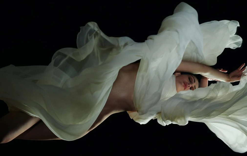 Woman draped in flowing white fabric against a black background, creating an ethereal and artistic visual with soft lighting.