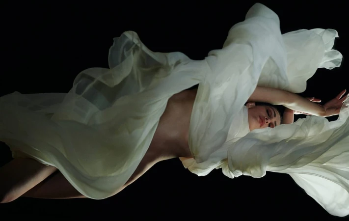 Woman draped in flowing white fabric against a black background, creating an ethereal and artistic visual with soft lighting.