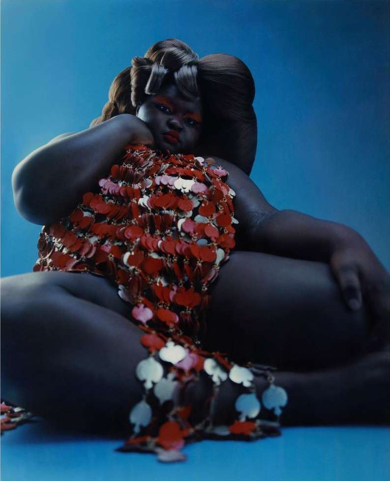 Portrait of an African American woman with artistic makeup and hairstyle wearing a red beaded top against a blue background in studio lighting.