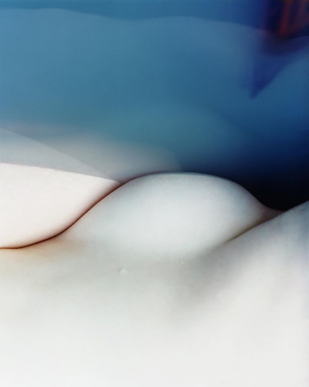 Close-up of a pale-skinned human torso with smooth skin and gentle curves, set against a blurred blue background suggesting water.