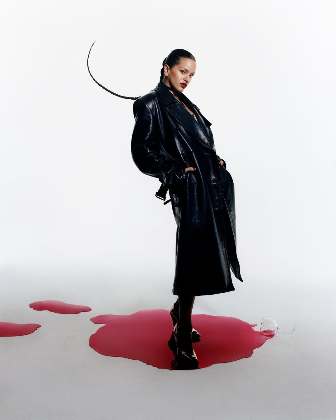 Fashion editorial shot of a stylish adult female in a black trench coat and boots posing indoors against a white background with spilled liquid.