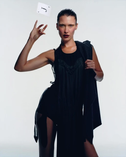 Elegant woman in black dress poses in photo studio, throwing playing card in the air, back jacket over the shoulder, dark lipstick.